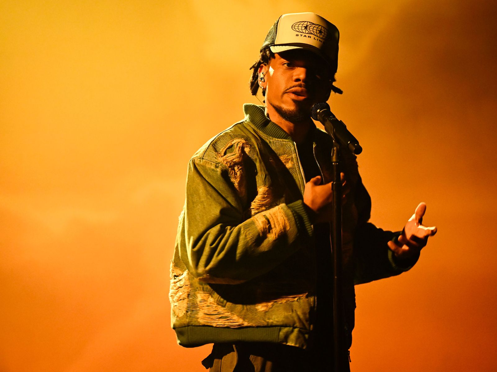 Chance the Rapper performs onstage wearing a cap and a jacket and holding a microphone against an orange background.
