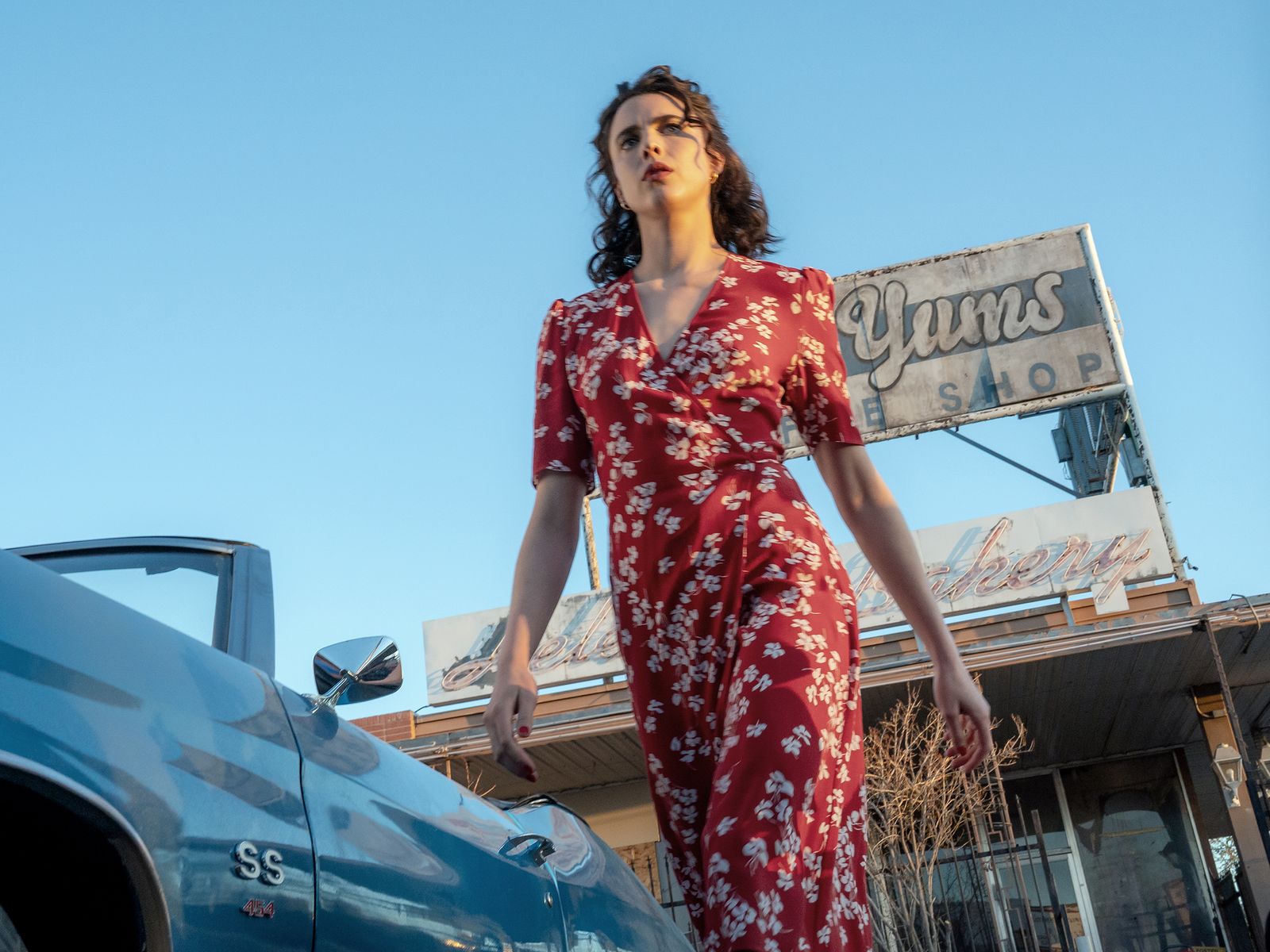 Shot from the ground shows woman wearing read dress with white flowers on it. She is mid walk and backed by a blue...