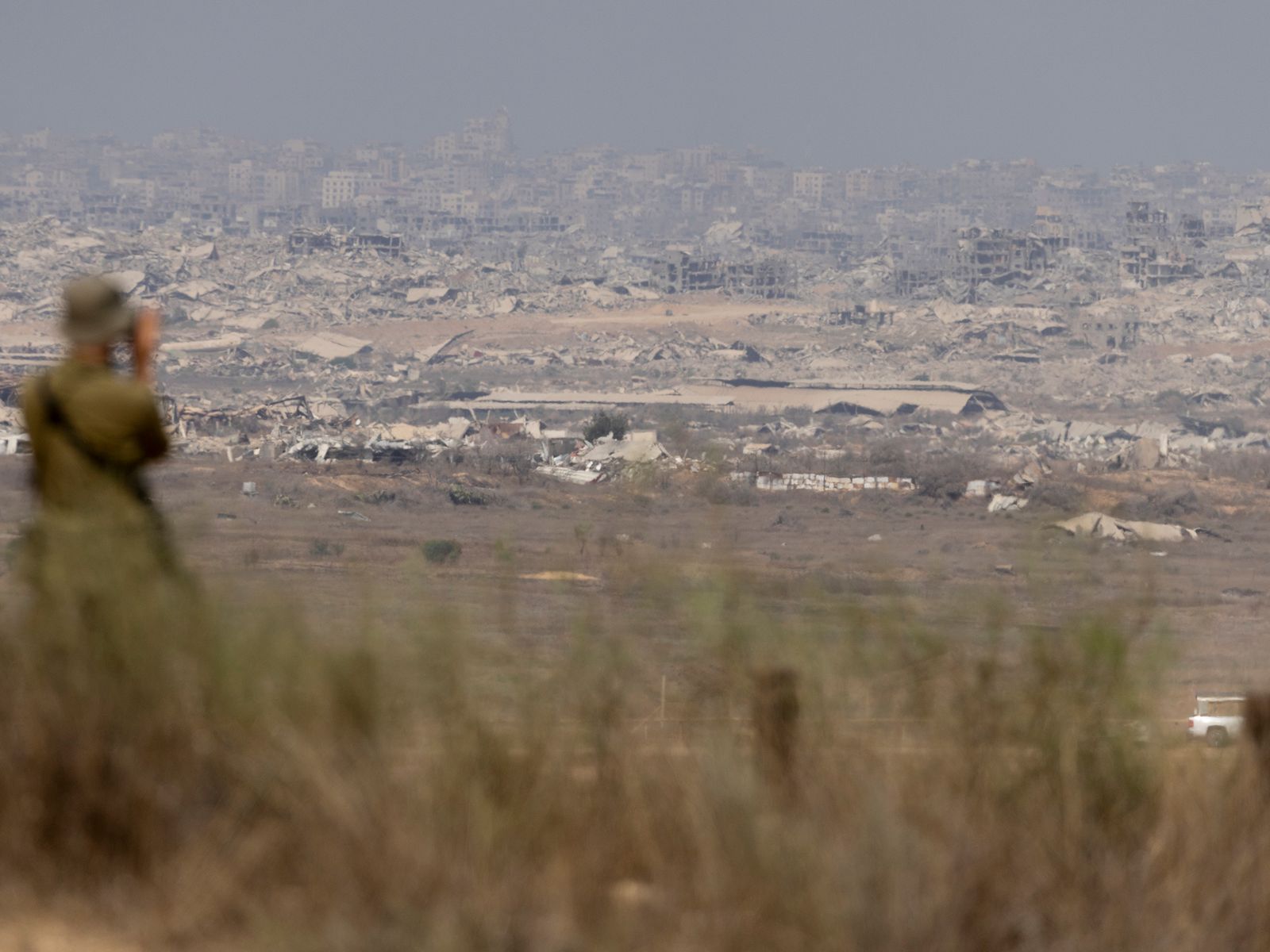 Blurred figure in soldier uniform stands amidst grass looking at the Northern Gaza Strip with binoculars. A destroyed...