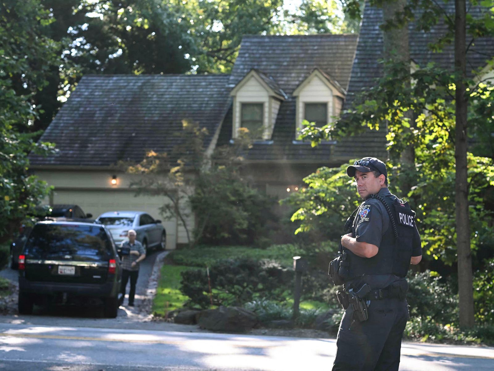 Police officer stands with back to camera looking over his shoulder directly at camera. Behind him a house and greenery...